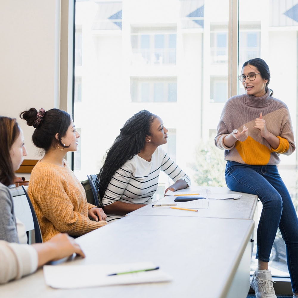 Female teacher sitting on desk speaking to three female students